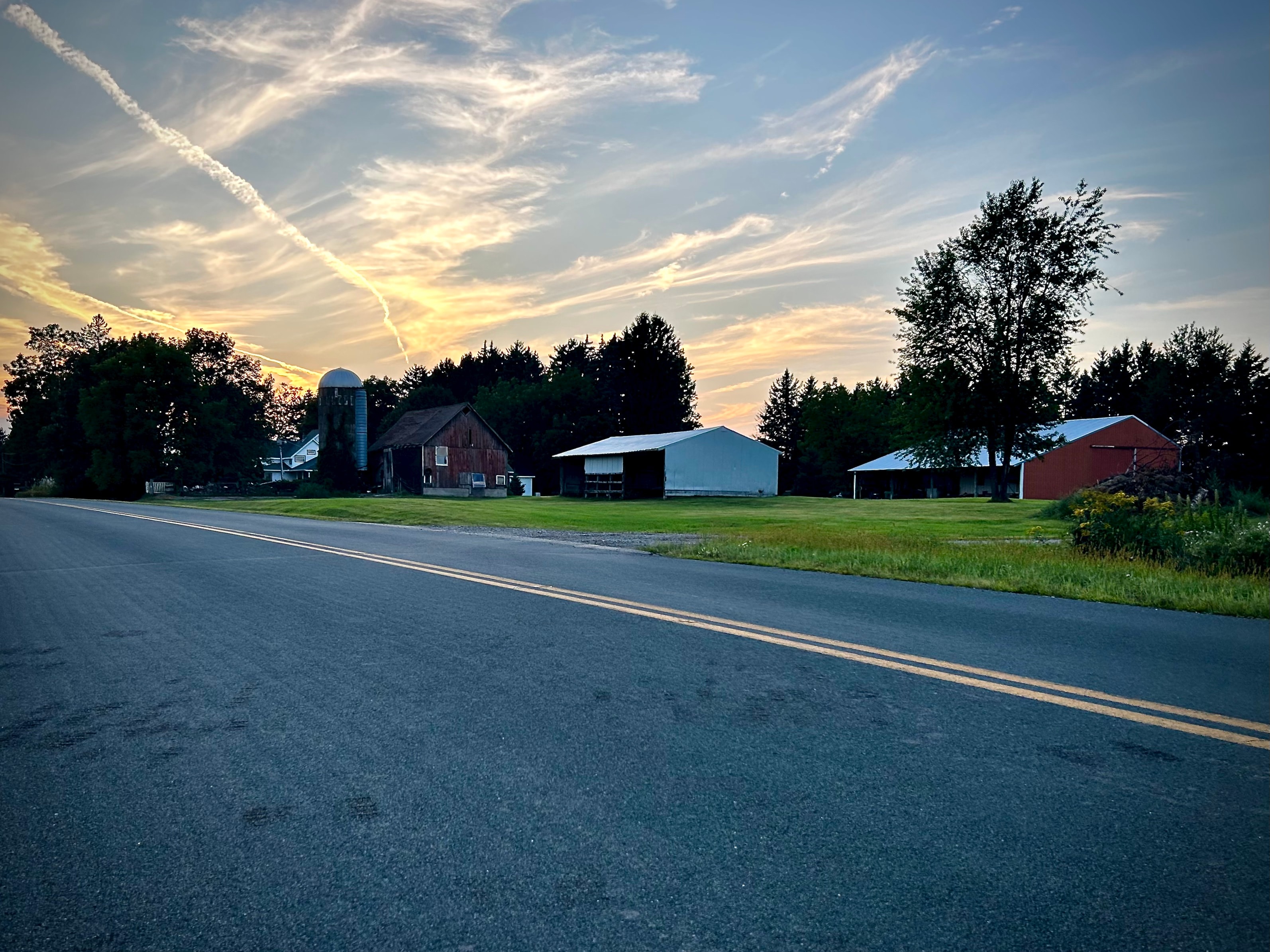 Rural road at sunset, farm barns and silo silhouetted against a warm evening sky